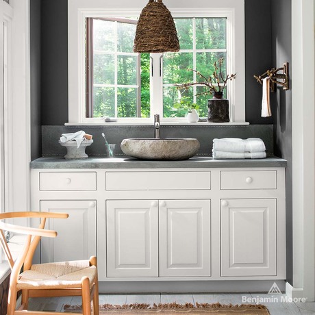 A white bathroom vanity with stone vessel sink underneath a charcoal gray wall with window. A white bathroom vanity with stone vessel sink underneath a charcoal gray wall with window.