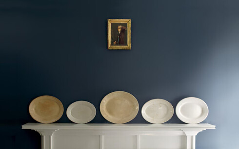 A white wood mantel features decorative plates, against a blue wall; a small antique painting above.
