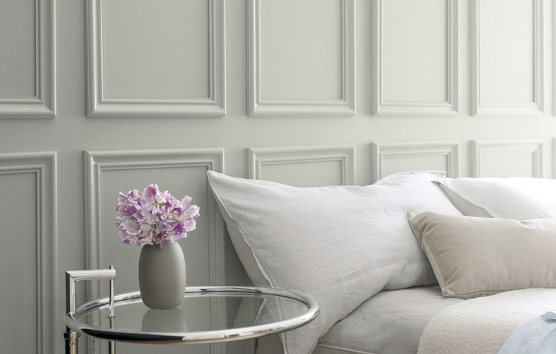 Gray wall with gray bedding and a glass side table featuring a small vase of purple flowers. Gray wall with gray bedding and a glass side table featuring a small vase of purple flowers.
