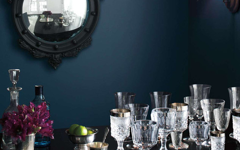 A dark blue-painted room with black framed mirror and crystal glasses on a table.